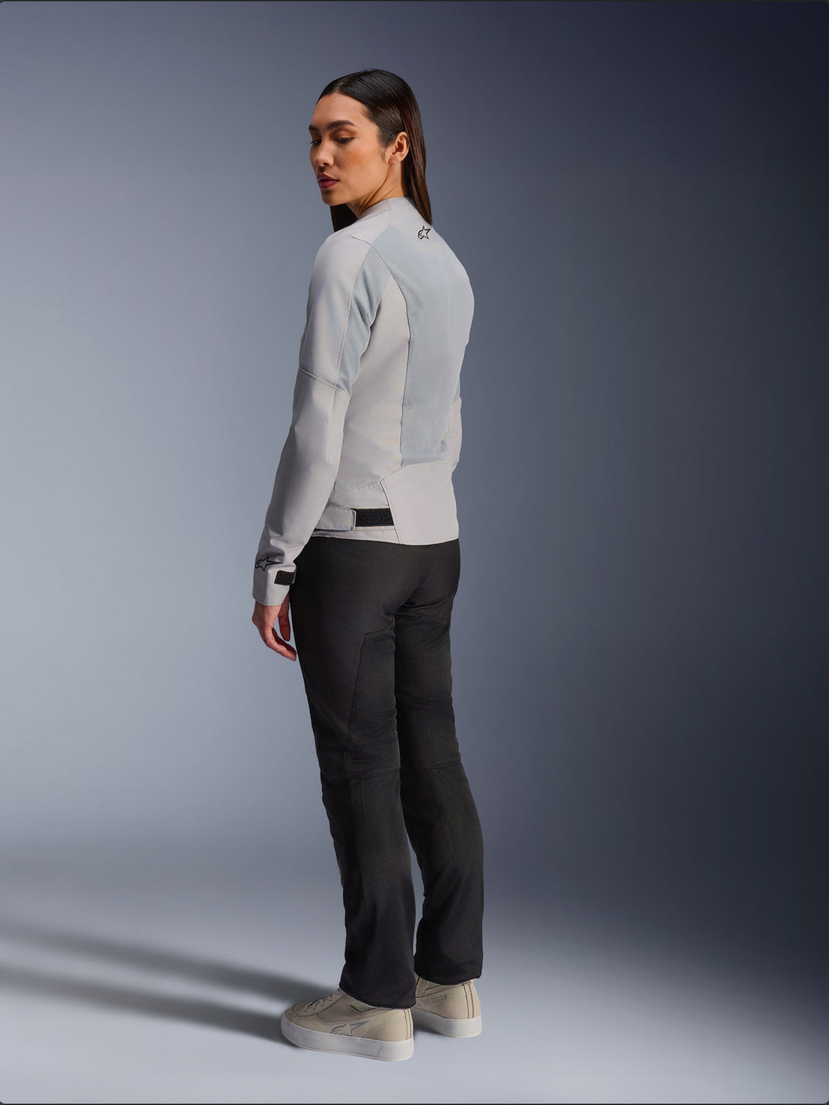 Mulher a usar o casaco Alpinestars Stella C-1 Air em prateado, vista traseira e lateral mostrando painéis de malha técnica e logótipo refletor no ombro, combinado com calças de motociclismo pretas e sapatilhas bege, estética de condução urbana contemporânea