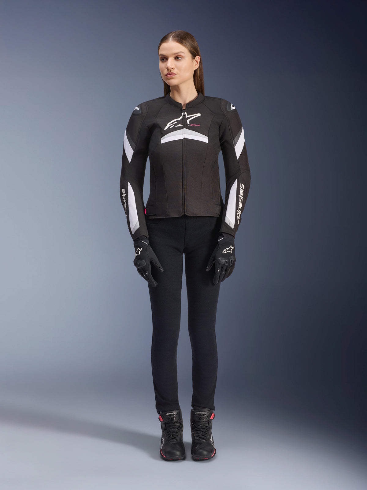 Modelo feminina a usar a Jaqueta Stella T-GP Plus R V4 da Alpinestars em preto e branco, jaqueta de motociclismo desportiva em tecido, combinada com calças, luvas e botas pretas, vista frontal com logótipo Alpinestars branco e detalhes nas mangas.