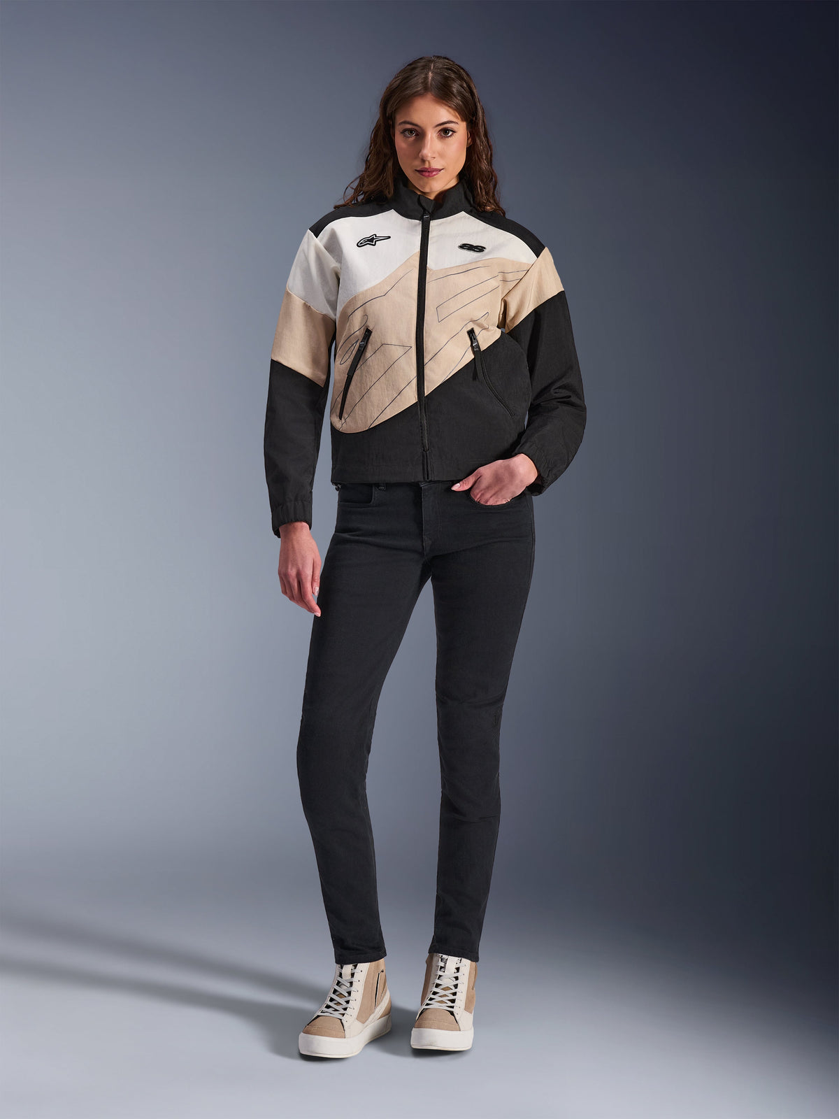 Casaco Alpinestars Stella Moshell para Mulher, casaco de motociclismo urbano com design assimétrico em blocos de cor preto, sand e off-white, combinado com calças pretas slim-fit e sapatilhas de cano alto beges, estética streetwear moderna para motociclistas.