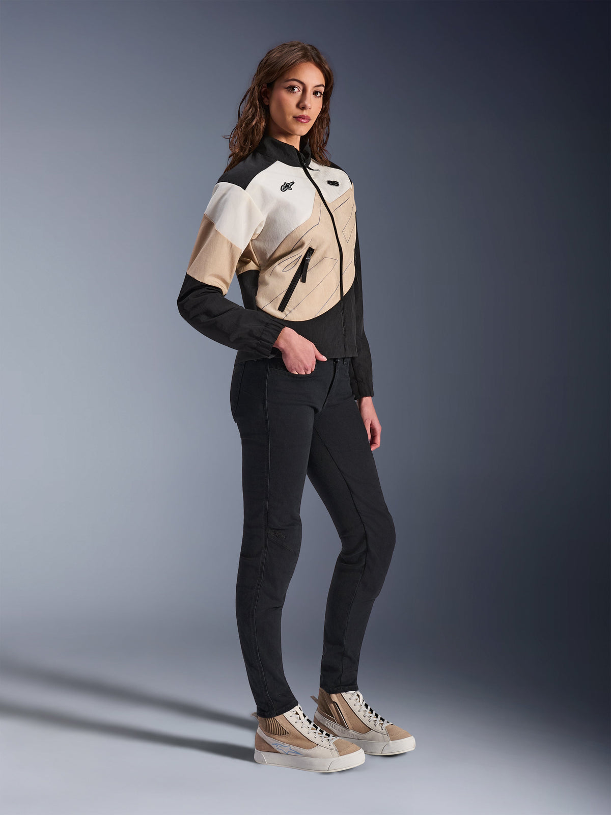 Casaco Alpinestars Stella Moshell, casaco urbano de motociclismo para mulher, preto, areia e off-white, com um design geométrico assimétrico, usado com calças pretas justas e sapatilhas beges, fotografia de estúdio de corpo inteiro de equipamento urbano contemporâneo.