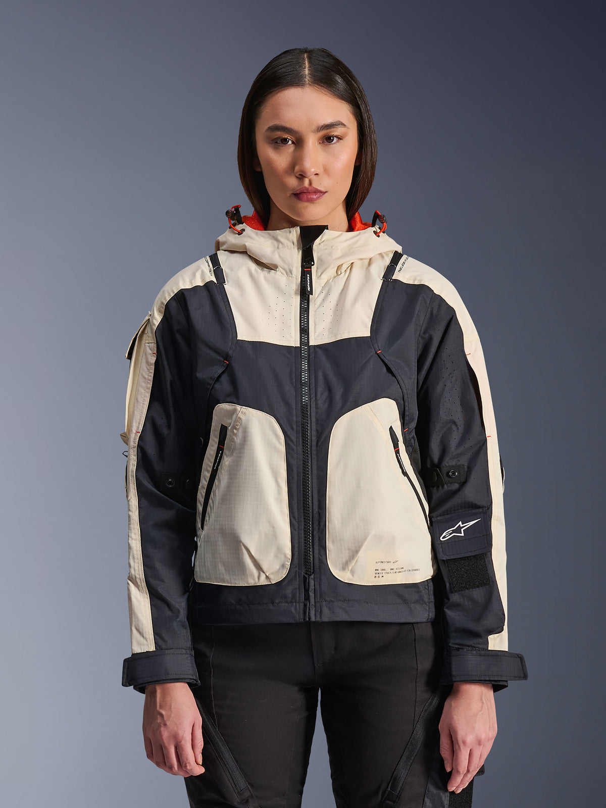 Alpinestars Molly Air Jacket, casaco de moto urbano para mulher, cor Black Almond, vista frontal numa modelo, com painéis ripstop em creme e preto, capuz com forro cor de laranja, fecho de correr frontal e grandes bolsos utilitários, equipamento de motociclismo contemporâneo inspirado no estilo streetwear