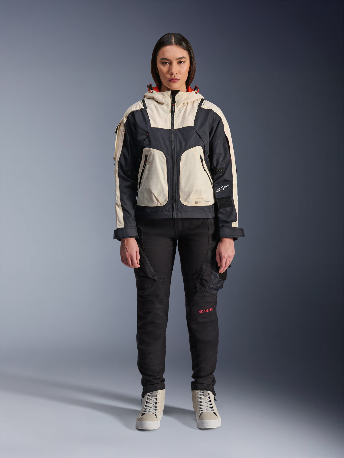Modelo feminina a usar o casaco Alpinestars Molly Air em Black Almond, com um design de blocos de cor com painéis em creme e preto e um capuz com forro vermelho, combinado com calças de motociclismo pretas e ténis creme, fotografia de estúdio de equipamento de condução urbano contemporâneo.