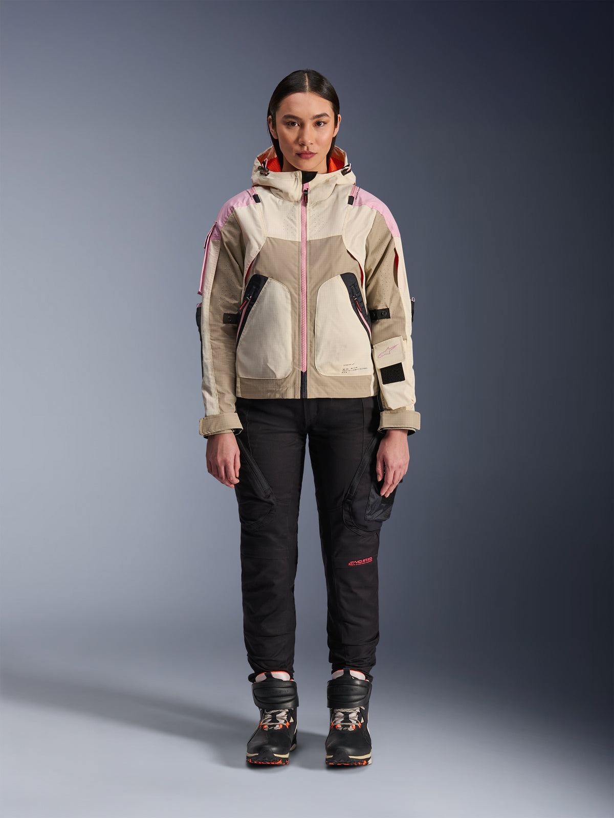 Modelo feminina a usar o Casaco Alpinestars Molly Air em Sand Pink Almond, acompanhado por calças técnicas de motociclismo pretas e botas, vista frontal de corpo inteiro, casaco de motociclismo urbano com design de inspiração utilitária e detalhes em cor-de-rosa