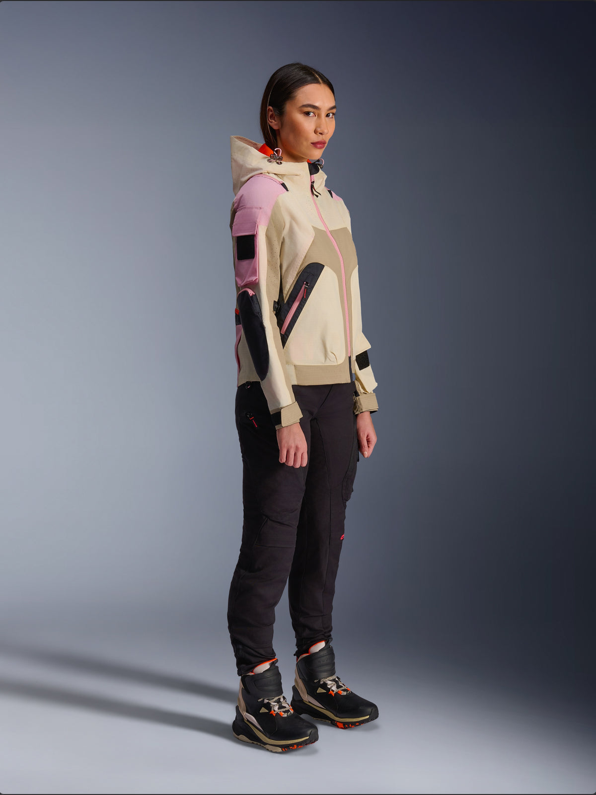 Modelo feminina a usar o Casaco Alpinestars Molly Air em Sand Pink Almond, combinado com calças técnicas escuras e botas de motociclismo de cano alto, casaco de motociclismo urbano com capuz e painéis multicoloridos, design contemporâneo inspirado em streetwear