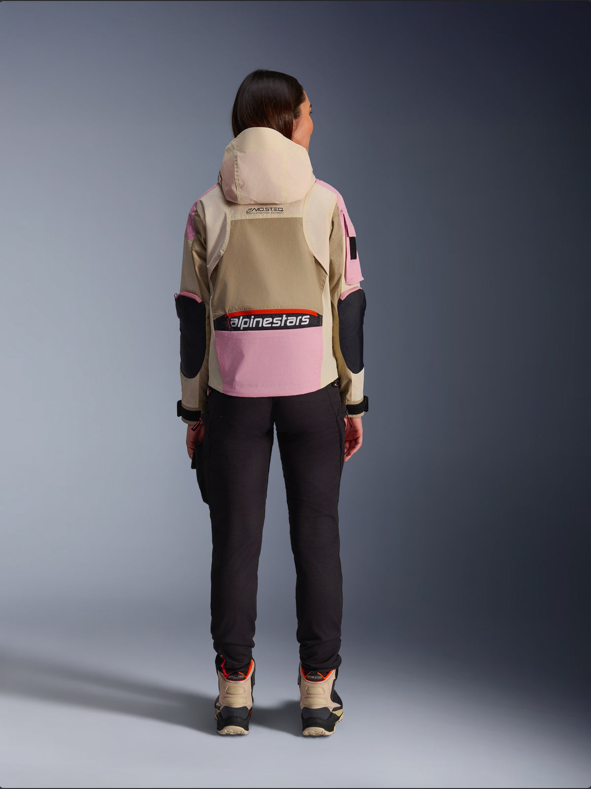 Motociclista feminina vista de costas a usar o casaco Alpinestars Molly Air em Sand Pink Almond, com capuz bege e painéis cor-de-rosa com pormenores pretos, combinado com calças de condução pretas e botas de motociclismo beges, vestuário de motociclismo urbano contemporâneo.