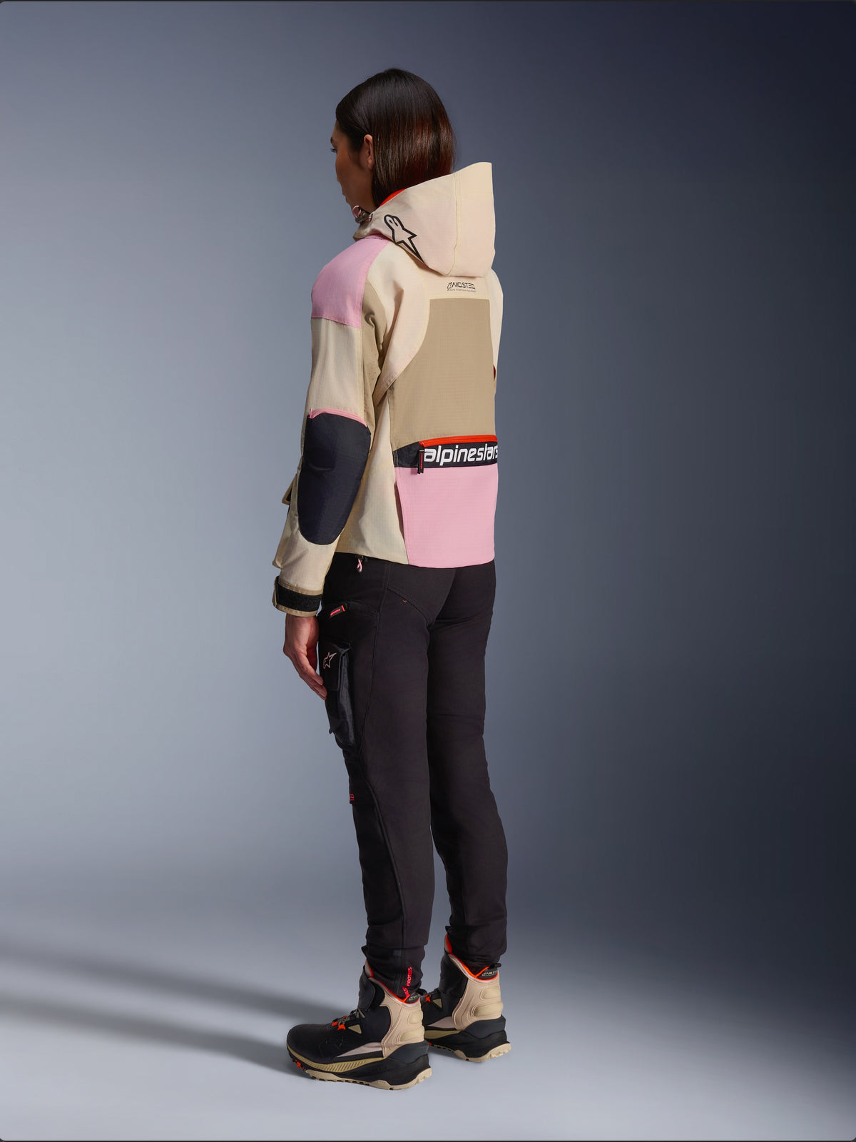 Motociclista feminina a usar o Casaco Alpinestars Molly Air em Sand Pink Almond, com um design em tons de bege e rosa, vista de perfil traseiro com calças de motociclismo pretas e botas técnicas a condizer, estilo de motociclismo urbano com capuz.