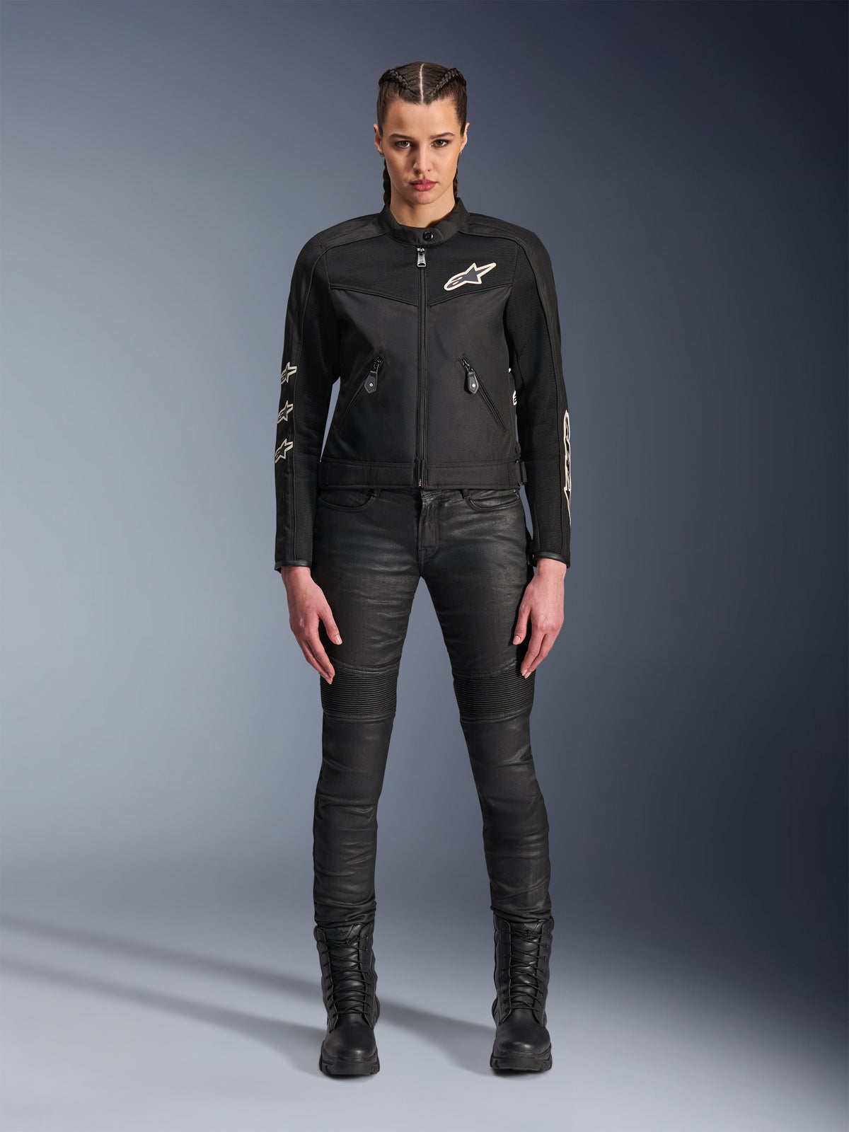 Modelo feminina a usar o casaco Alpinestars Stella T-Dyno Air, casaco de motociclismo para mulher em preto, com painéis de malha 3D e logótipos Astars brancos, combinado com calças de condução pretas de corte justo e botas pretas com atacadores, estilo de equipamento de condução urbana.