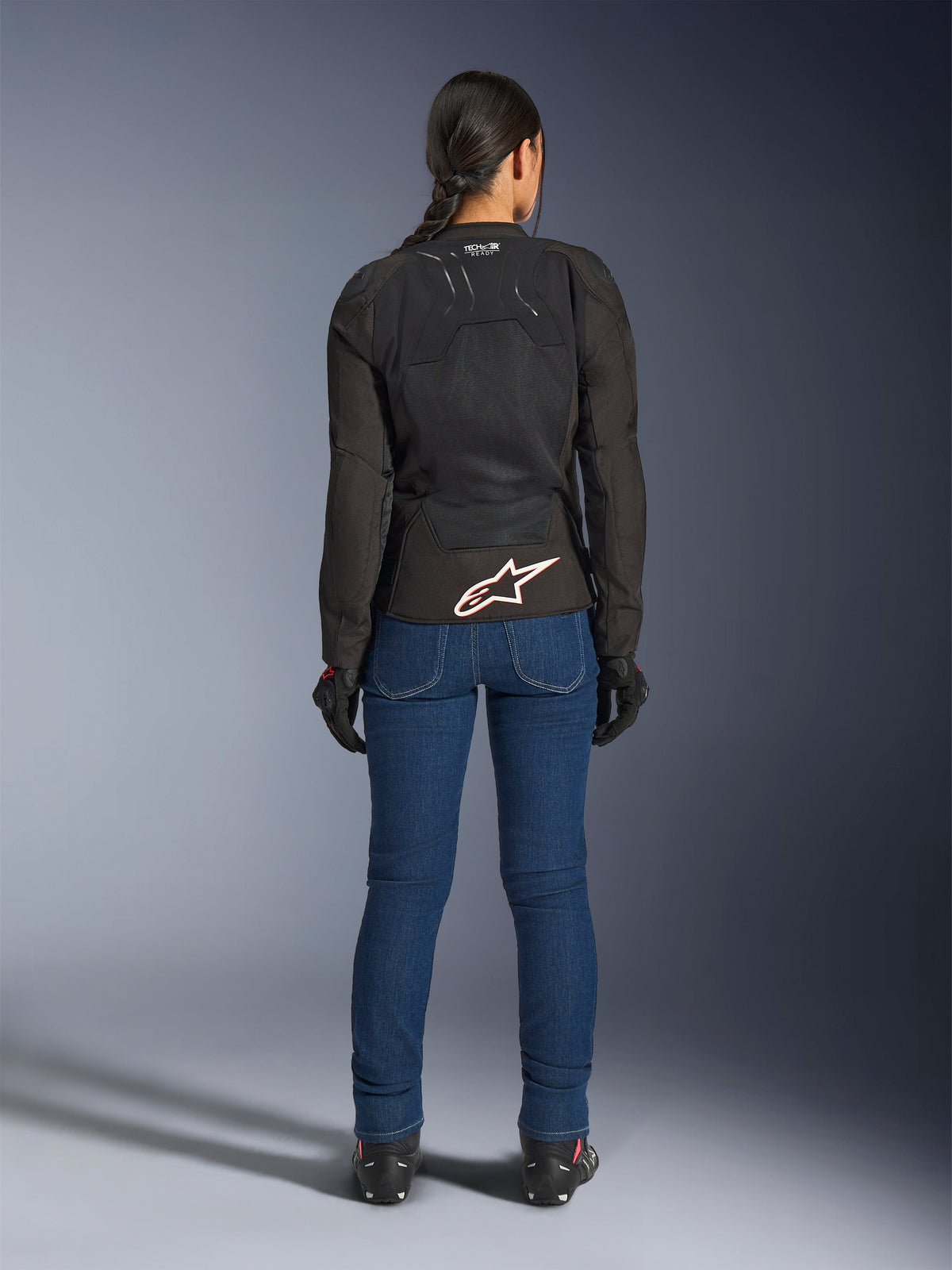 Condutora vista de costas a usar o Casaco Alpinestars Stella SMX Air em preto, combinado com calças de ganga azuis para motociclismo e luvas pretas, com design têxtil ventilado, logótipo de estrela branca na zona lombar e marca Tech-Air Ready, concebido para condução desportiva em climas quentes
