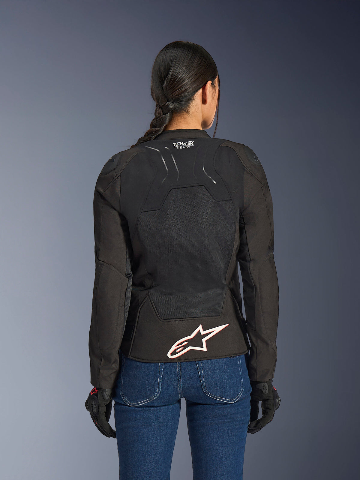 Casaco Alpinestars Stella SMX Air, vista traseira numa modelo feminina, têxtil preto com painéis de malha de alto fluxo, marcação 'TECH-AIR READY' na parte superior das costas, logótipo Alpinestars grande em branco com contorno vermelho na parte inferior das costas, combinado com calças de ganga azuis e luvas de condução pretas