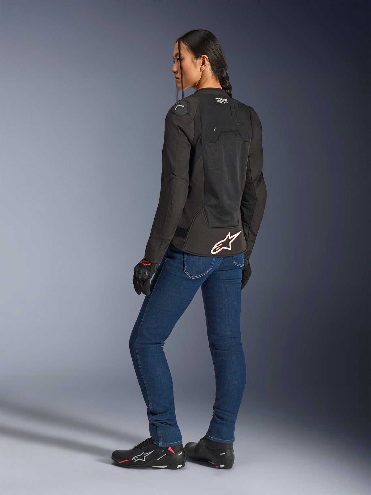 Casaco Alpinestars Stella SMX Air, preto com logótipo branco e vermelho, usado por uma motociclista numa vista de três quartos traseira, combinado com calças de ganga de motociclismo azuis e calçado de condução preto, apresentando uma construção em malha técnica para condução desportiva em clima quente.