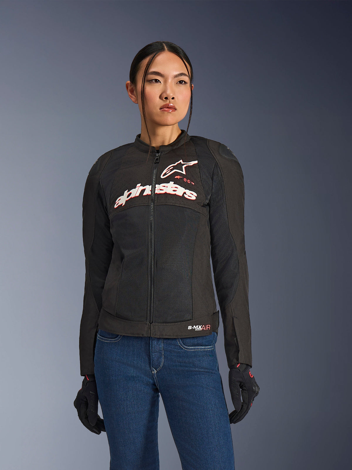 Mulher a usar o Casaco Alpinestars Stella SMX Air, preto, branco e vermelho fluo, casaco têxtil de motociclismo com malha ventilada e logótipo branco no peito, combinado com calças de ganga azuis e luvas de condução pretas, corte feminino para condução desportiva