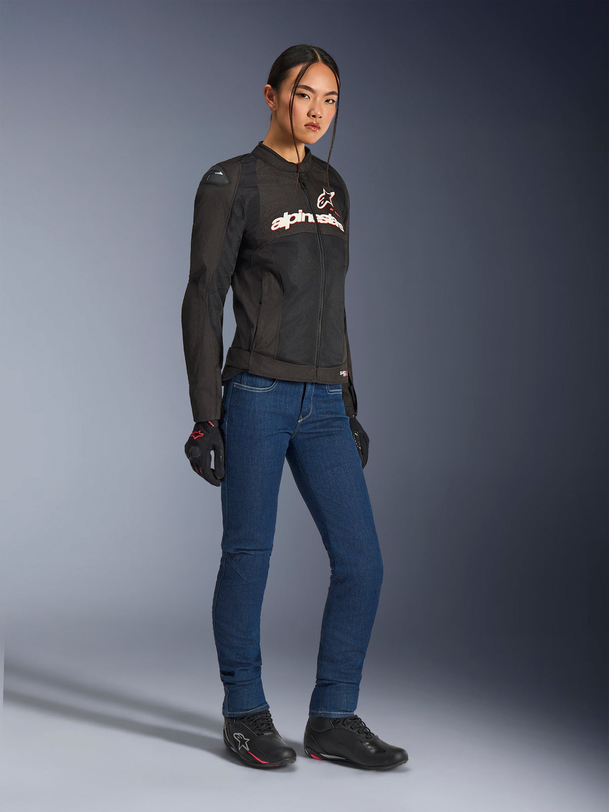 Alpinestars Stella SMX Air Jacket, casaco de motociclismo têxtil para mulher, preto com detalhes em branco e vermelho, usado com calças de ganga de motociclismo azuis, luvas pretas e sapatos de motociclismo pretos, equipamento de condução urbano e desportivo para mulher.