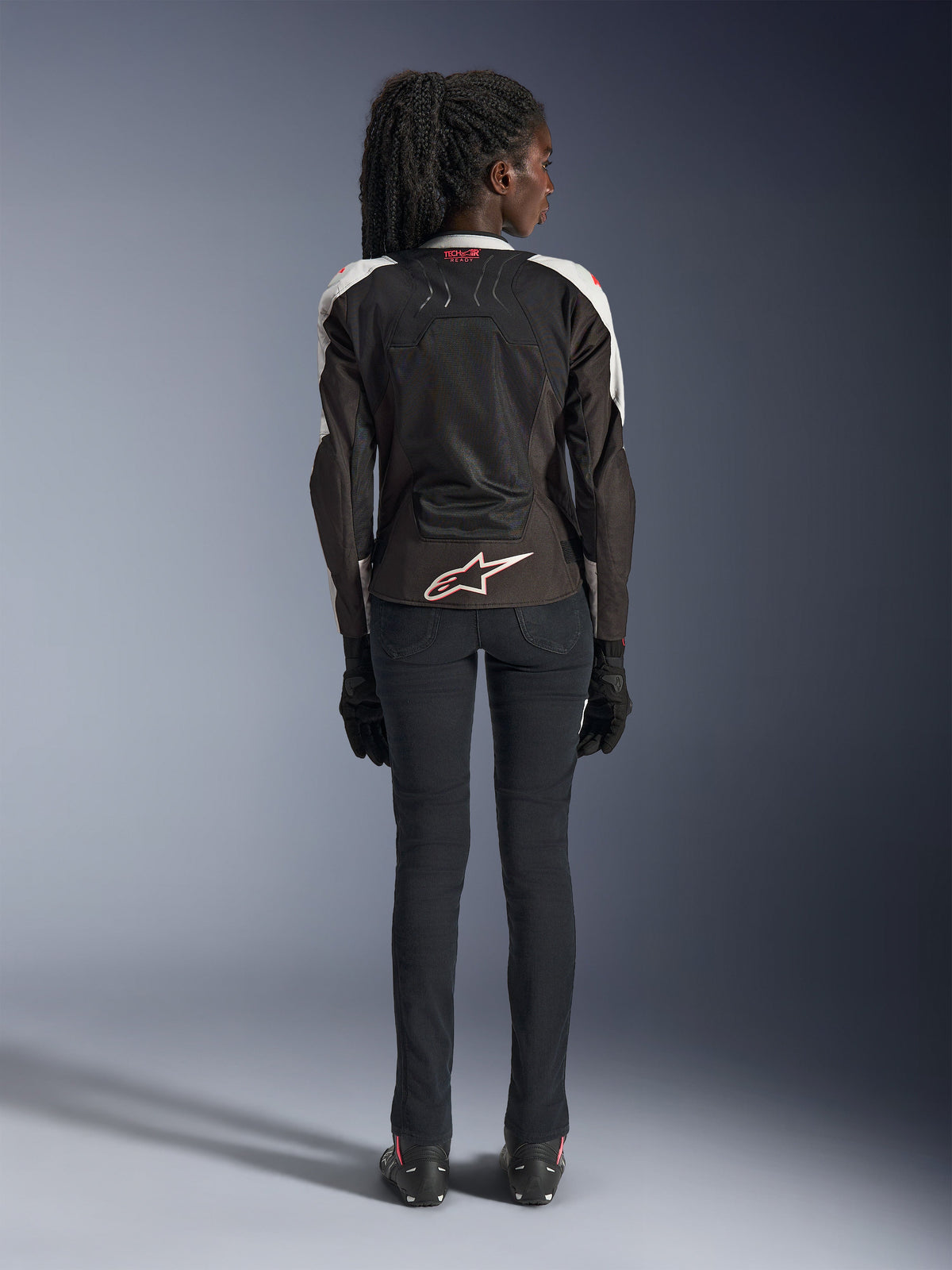 Casaco Alpinestars Stella SMX Air para Mulher, Ice Gray Black Coral Fluo, vista traseira com painéis de malha ventilada, logótipo Alpinestars branco na parte inferior das costas e marca Tech-Air Ready na gola, usado com calças e luvas de motociclismo pretas.