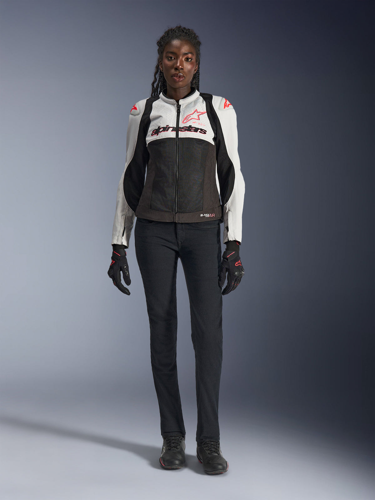 Casaco Alpinestars Stella SMX Air, Ice Gray, Black e Coral Fluo, usado por uma modelo feminina com calças de motociclismo pretas e luvas, com estrutura em malha e sliders de proteção nos ombros, equipamento de motociclismo desportivo para mulher
