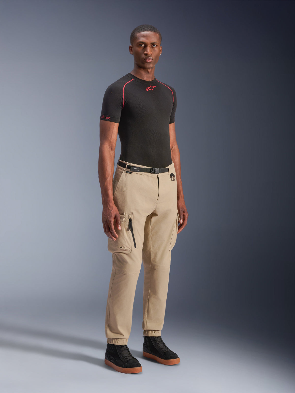 Alpinestars Flex-AST Cargo Pants - Short, calças de motociclismo urbanas, bege claro, vestidas por um modelo com uma camada de base preta e sapatilhas, com um corte regular tapered, bainhas elásticas e design cargo com múltiplos bolsos