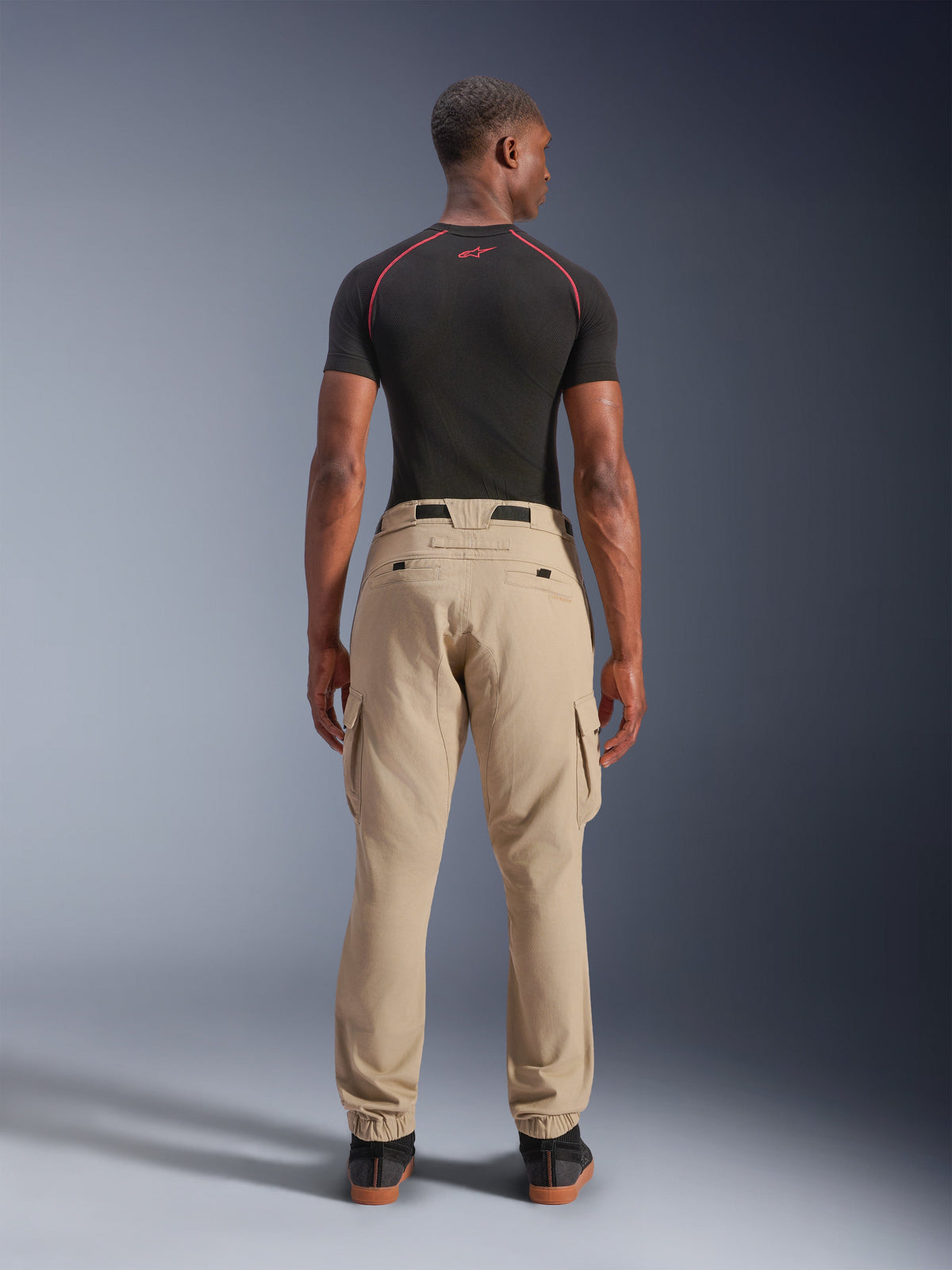 Alpinestars Flex-AST Cargo Pants - Short, bege claro, vista traseira num modelo, corte regular afunilado com bainhas elásticas e bolsos cargo, usado com uma base layer técnica preta com detalhes vermelhos, vestuário de motociclismo urbano moderno