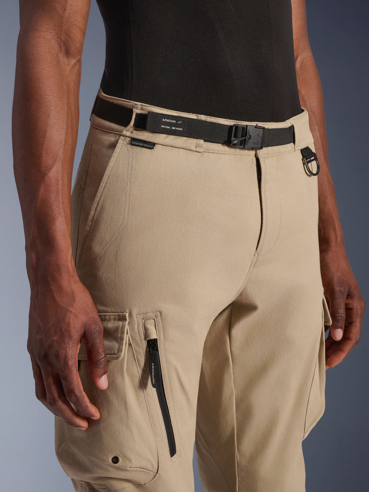 Alpinestars Flex-AST Cargo Pants - Short, bege claro, vista aproximada da cintura e coxa, com cinto ajustável preto com a marca Alpinestars, anel em D metálico e bolsos cargo funcionais com fechos de correr e abas com botões de pressão, equipamento de motociclismo urbano contemporâneo