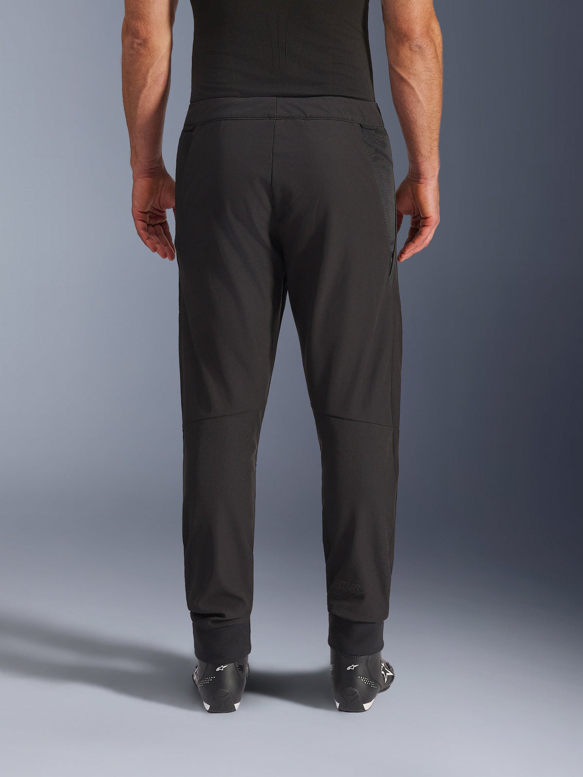 Calças Alpinestars Aeroshell Airflow, pretas, vista traseira, com zonas perfuradas a laser nas panturrilhas e painéis de malha, usadas com botas pretas Alpinestars, design softshell leve para desporto e turismo