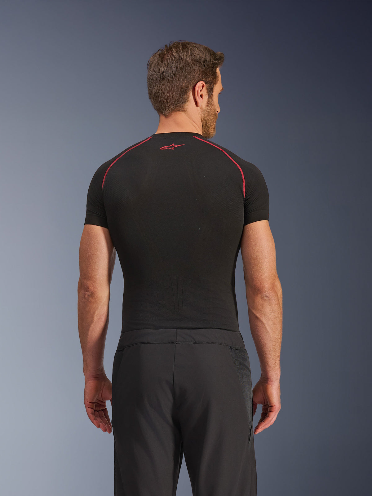 Vista traseira de um modelo a usar Calças Alpinestars Aeroshell Airflow, pretas, construção leve em softshell com painéis laterais em malha integrados, combinadas com uma camada de base preta com costuras de contraste vermelhas, vestuário desportivo para motociclista