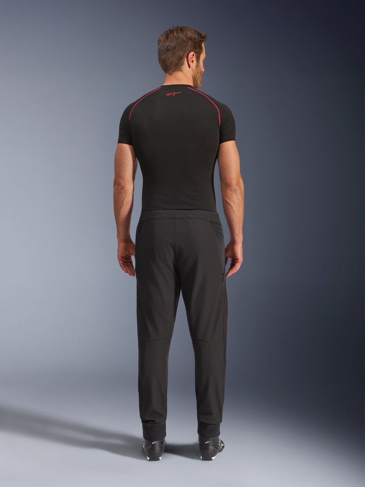 Alpinestars Aeroshell Airflow Pants, calças de motociclismo pretas, vista traseira mostrada num modelo com camada de base preta e botas, com um chassis leve de softshell e painéis de malha integrados para ventilação.