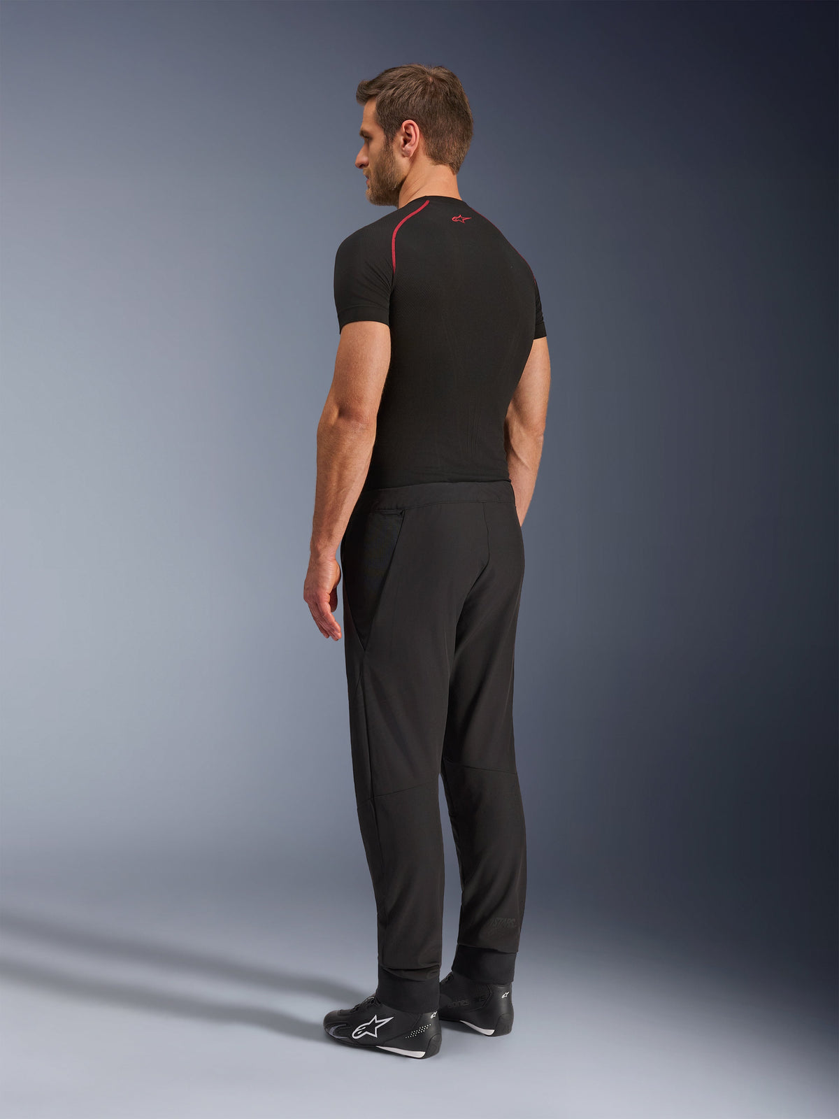 Alpinestars Aeroshell Airflow Pants, calças de motociclismo pretas, tecido softshell leve, usadas com uma t-shirt técnica de manga curta preta e botas de condução pretas, vista de perfil lateral traseira mostrando o corte justo e a construção ergonómica