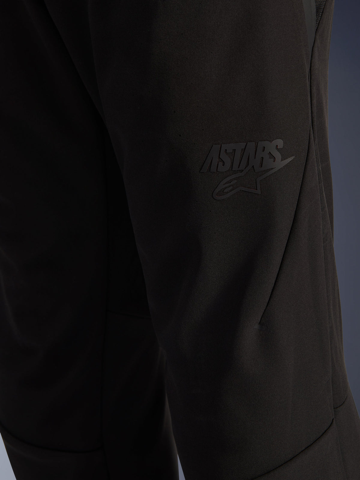 Alpinestars Aeroshell Airflow Pants, calças de motociclismo pretas, vista de pormenor da zona da coxa com logótipo ASTARS preto em relevo, material softshell elástico leve, design de costuras ergonómico