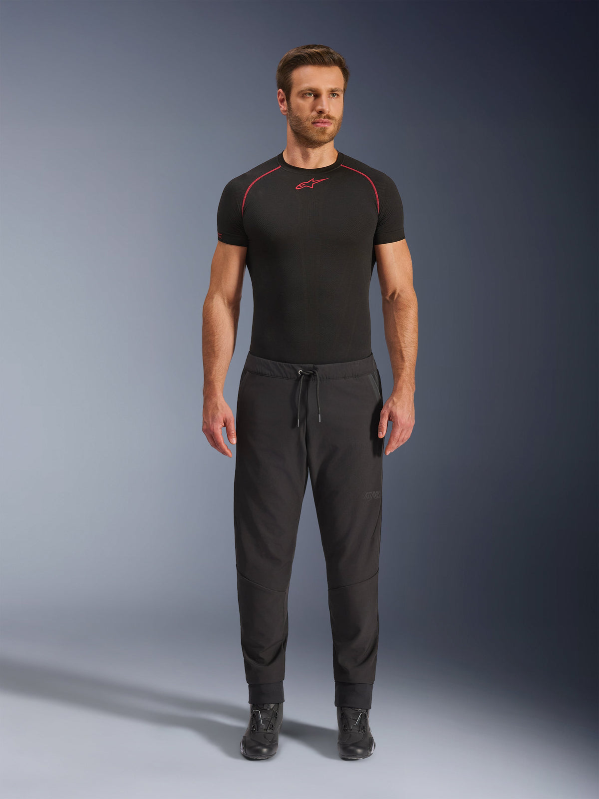 Modelo a usar calças Alpinestars Aeroshell Airflow em preto, combinadas com uma camada de base preta de manga curta e botas de motociclismo pretas. Estas calças de motociclismo leves em softshell apresentam uma cintura com cordão e punhos elásticos, concebidas para motociclistas de desporto e turismo em condições de clima quente.