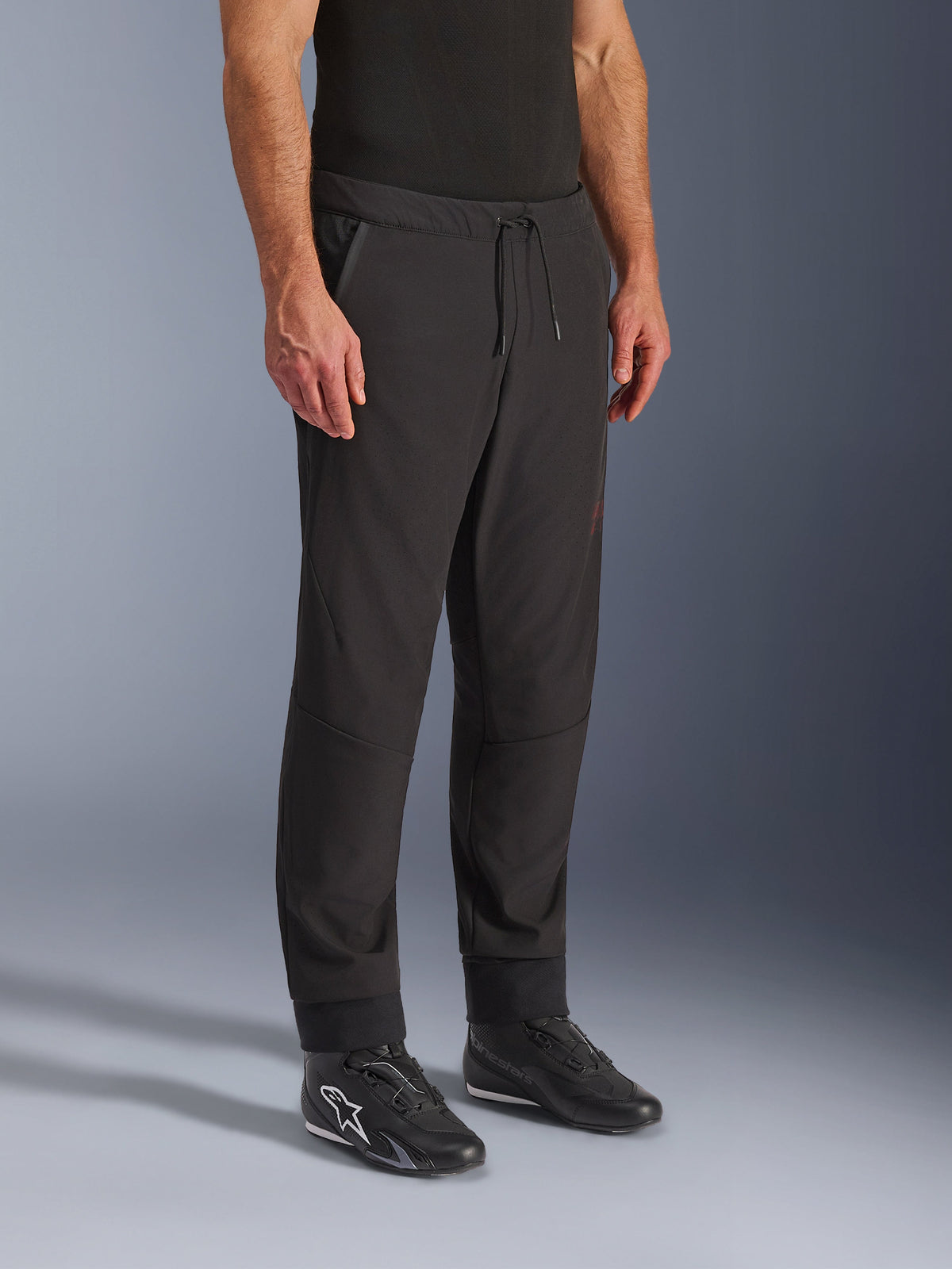 Alpinestars Aeroshell Airflow Pants, calças de motociclismo pretas, usadas por um modelo com calçado de motociclismo preto, com cintura com cordão, punhos nos tornozelos e tecido softshell perfurado a laser para máxima ventilação