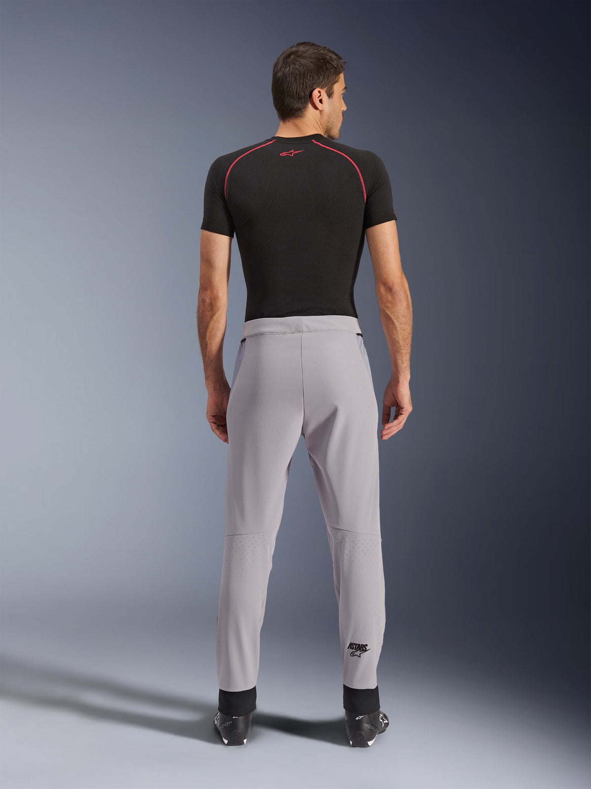 Alpinestars Aeroshell Airflow Pants, Cinzento Médio, vista traseira mostrando zonas perfuradas a laser nas panturrilhas, construção em softshell elástico leve, usadas com camisola preta de manga curta e botas de motociclista, design sport-fit para condução em climas quentes