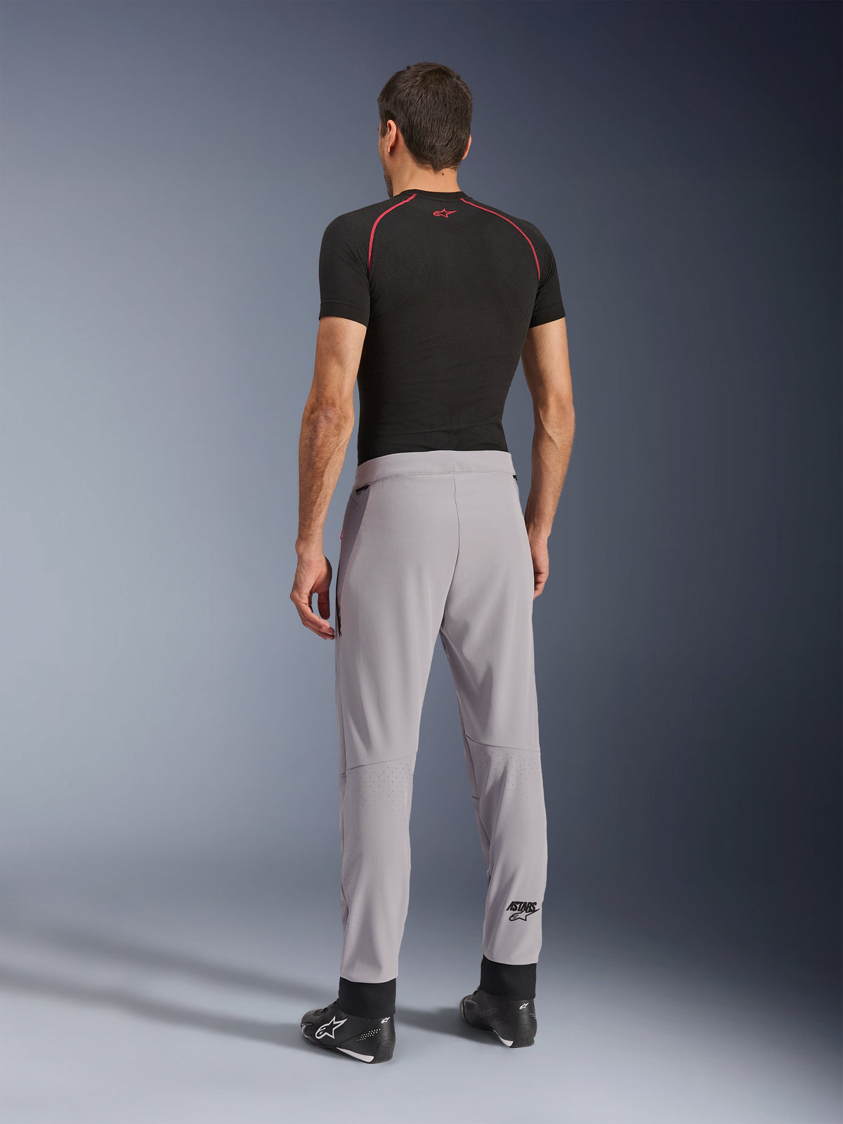 Vista traseira de um modelo a usar as calças Alpinestars Aeroshell Airflow em Mid Gray, combinadas com uma camisola de camada base preta e sapatos pretos, destacando as zonas perfuradas a laser nas panturrilhas e o corte desportivo atlético.