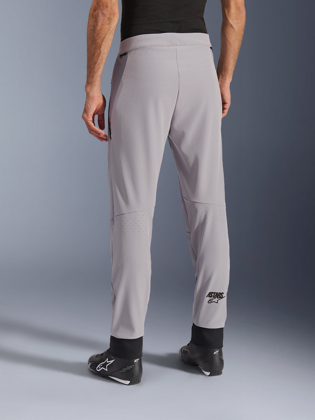 Calças Alpinestars Aeroshell Airflow, Cinza Médio, vista traseira mostrando zonas perfuradas a laser nas panturrilhas, logótipo ASTARS na parte inferior da perna, punhos pretos ajustáveis no tornozelo, usadas com botas de motociclista pretas, calças têxteis leves de condução desportiva