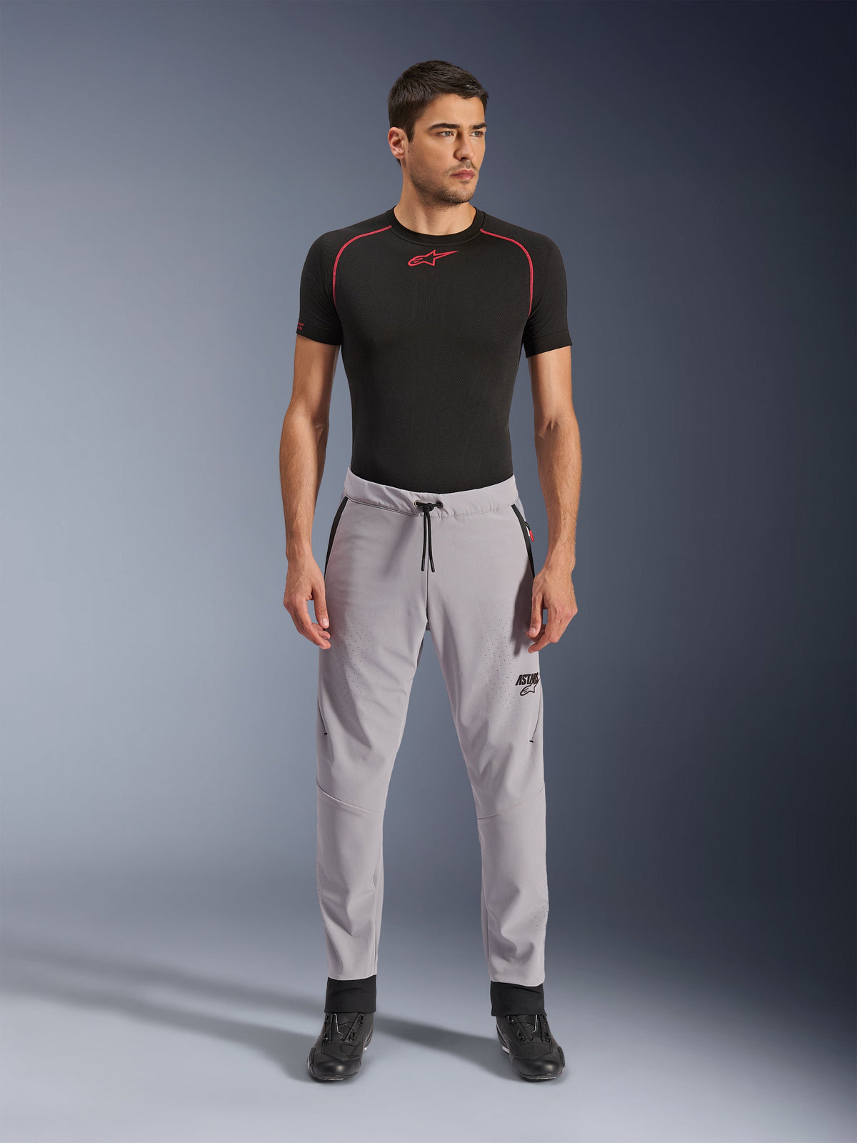 Calças Alpinestars Aeroshell Airflow, cinzento médio, calças têxteis leves com zonas de ventilação perfuradas a laser e cintura com cordão, vestidas por um modelo com camisola de compressão preta e botas de motociclismo pretas, design de condução desportiva limpo e moderno