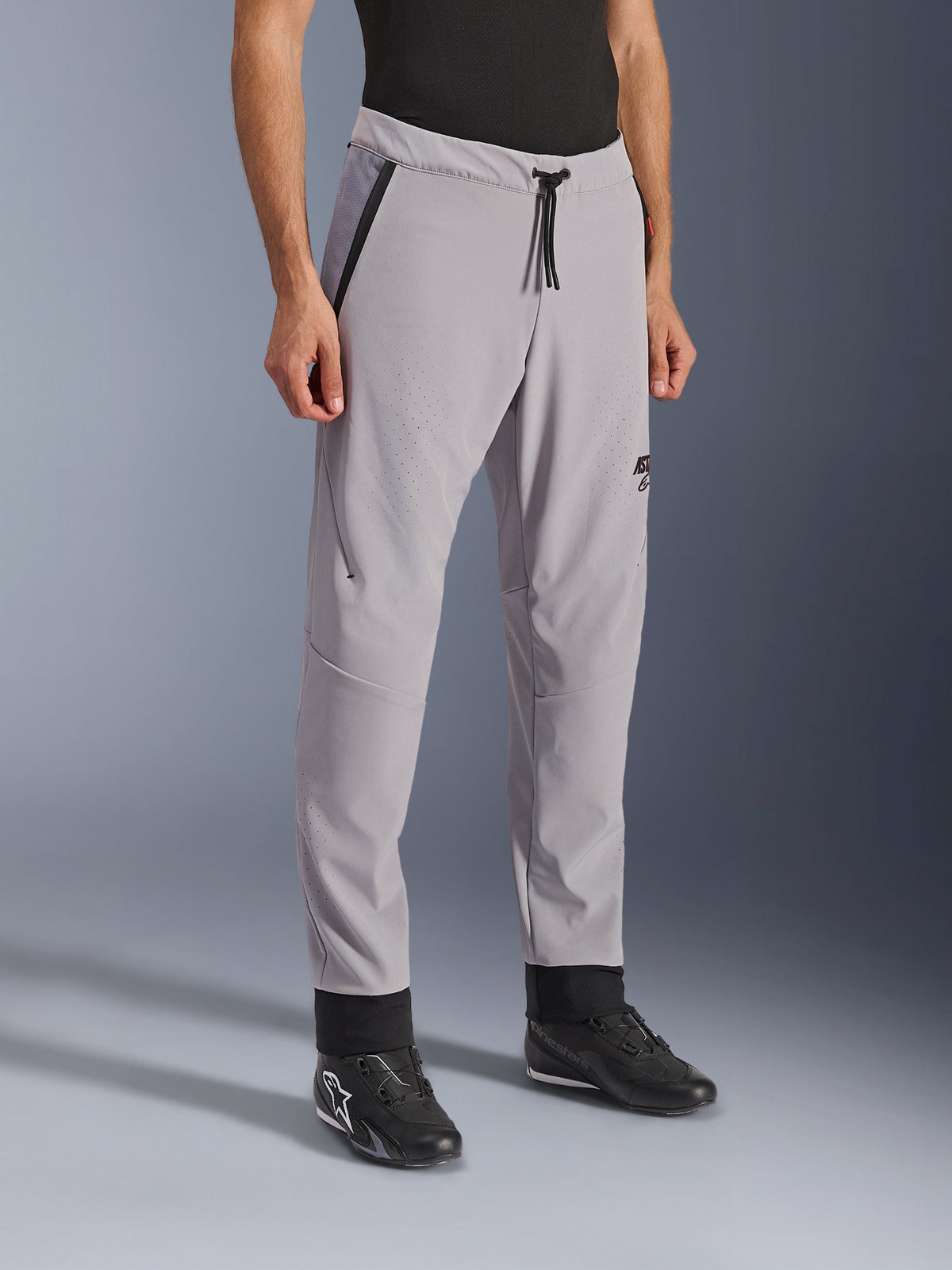 Alpinestars Aeroshell Airflow Pants, cinzento médio, calças de motociclismo leves com zonas de ventilação perfuradas a laser nas coxas e canelas, cintura elástica com cordão, corte desportivo justo, apresentadas num modelo com sapatos de condução pretos.
