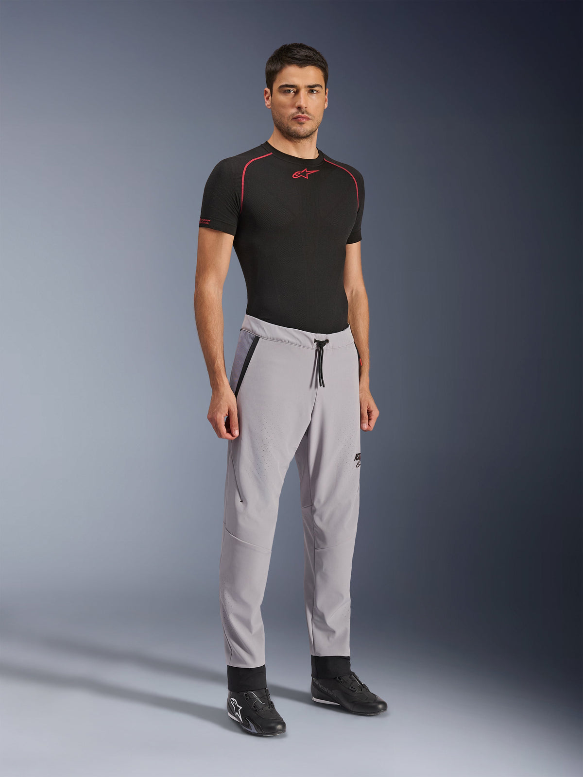 Calças Alpinestars Aeroshell Airflow, calças de motociclismo, cinzento médio, vestidas por um modelo com t-shirt técnica preta e sapatos de condução pretos, com zonas de ventilação perfuradas a laser, cintura elástica com cordão e corte desportivo aerodinâmico.