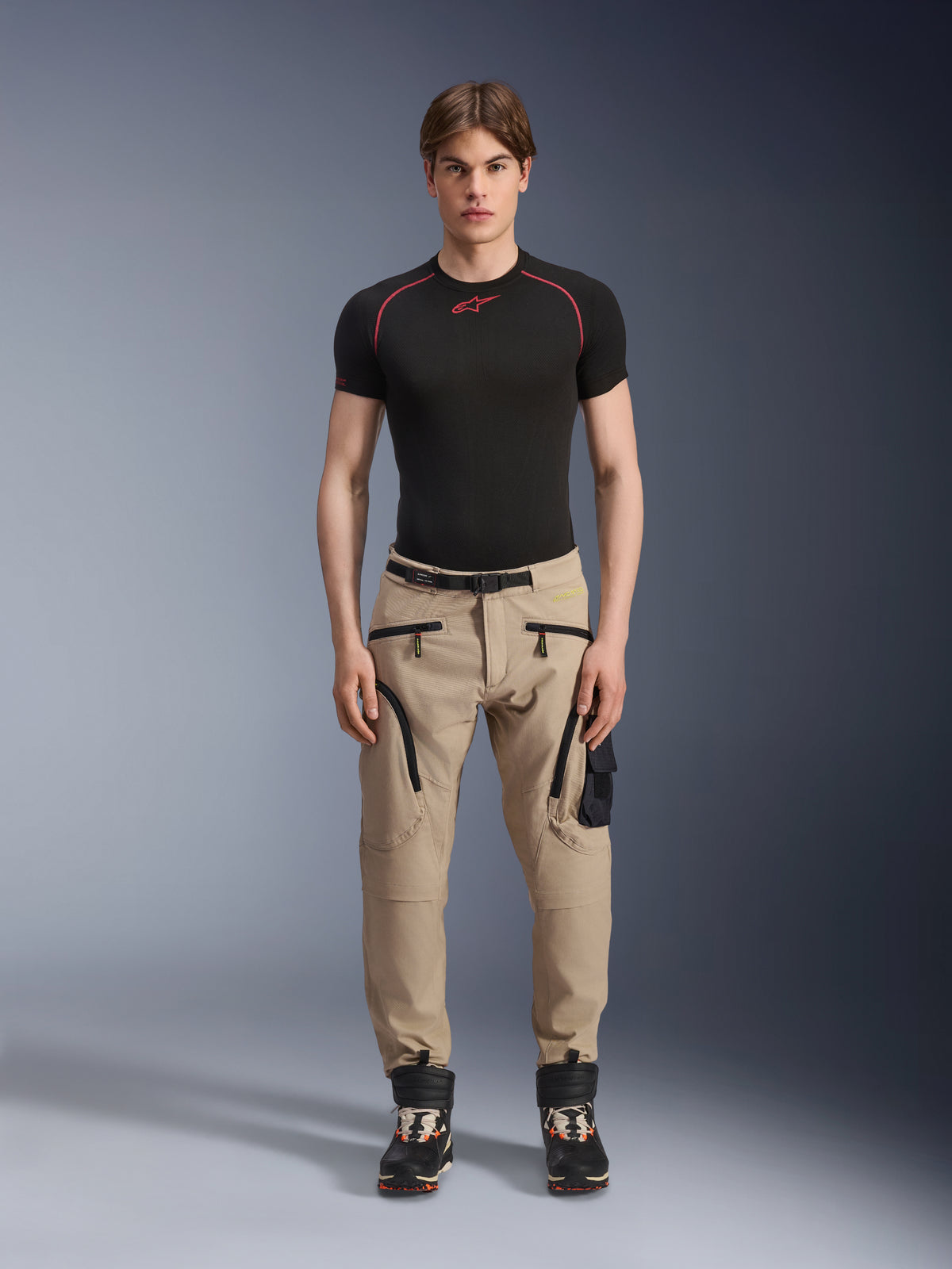 Modelo masculino a usar calças Alpinestars Acteon em cor Sand, combinadas com uma camada de base preta de manga curta e botas técnicas pretas, com bolsos cargo com fechos de correr pretos e um corte afunilado, equipamento de motociclismo urbano contemporâneo