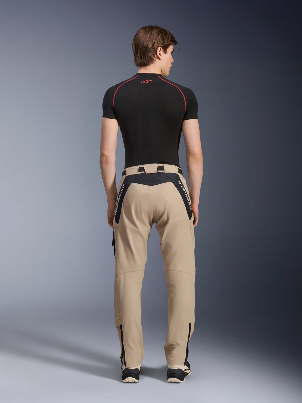 Calças Alpinestars Acteon em Sand, calças de motociclismo urbanas modernas vistas de costas, com painéis pretos e o logótipo Alpinestars a branco, corte afunilado com fechos de correr na bainha, usadas com uma camisola preta de manga curta e ténis.