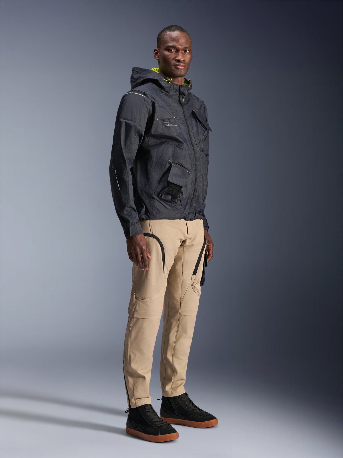 Calças Alpinestars Acteon, calças de motociclismo urbanas, cor Areia, vestidas por um modelo com um casaco técnico escuro e ténis pretos, com um corte afunilado, detalhes de fechos de correr pretos e bolsos tipo cargo, design contemporâneo inspirado no desporto motorizado