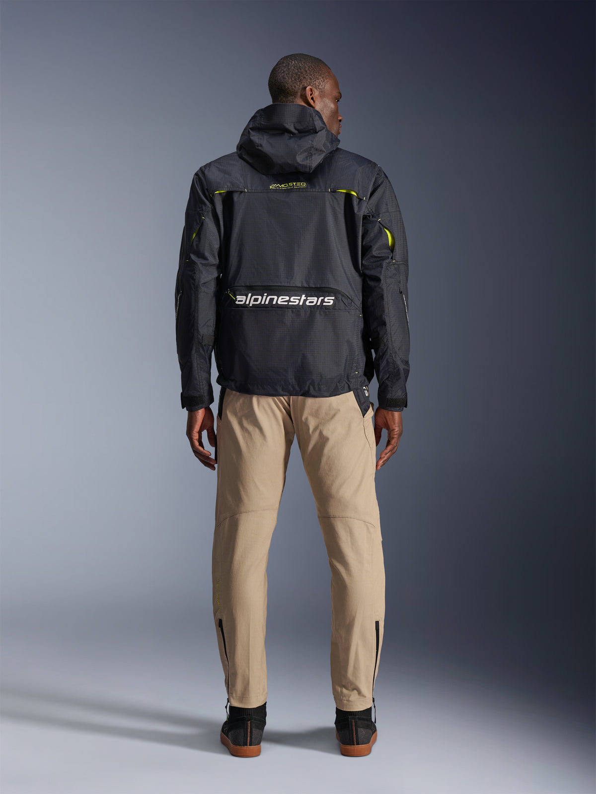 Calças Alpinestars Acteon, calças de motociclismo urbanas, cor areia, vistas de costas num modelo a usar um casaco técnico preto com capuz e botas escuras, apresentando uma perna afunilada com fechos de correr nos tornozelos e uma silhueta moderna e clean.