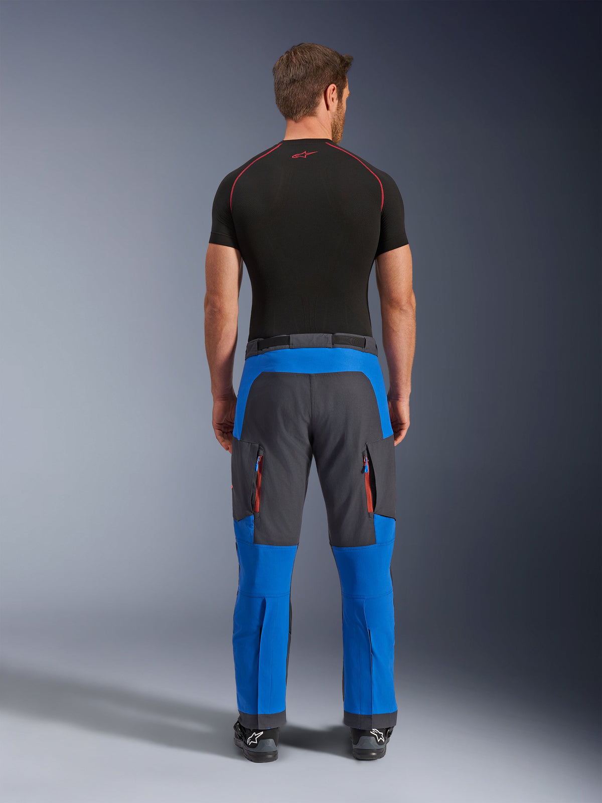 Alpinestars Maxdura Dual Pants em azul royal e cinzento escuro, calças de motociclismo de aventura vistas de trás, com bolsos cargo com fecho éclair e detalhes cor de laranja, design para usar por cima da bota, combinadas com uma camada base preta de manga curta