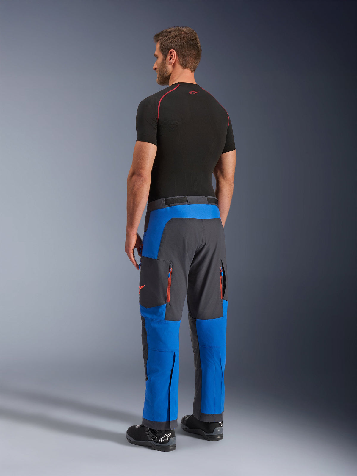Alpinestars Maxdura Dual Pants, calças de motociclismo de aventura, azul royal e cinzento escuro, vista traseira vestida por um modelo, com aberturas de ventilação com fecho cor de laranja, corte de aventura sobre a bota e material elástico durável.
