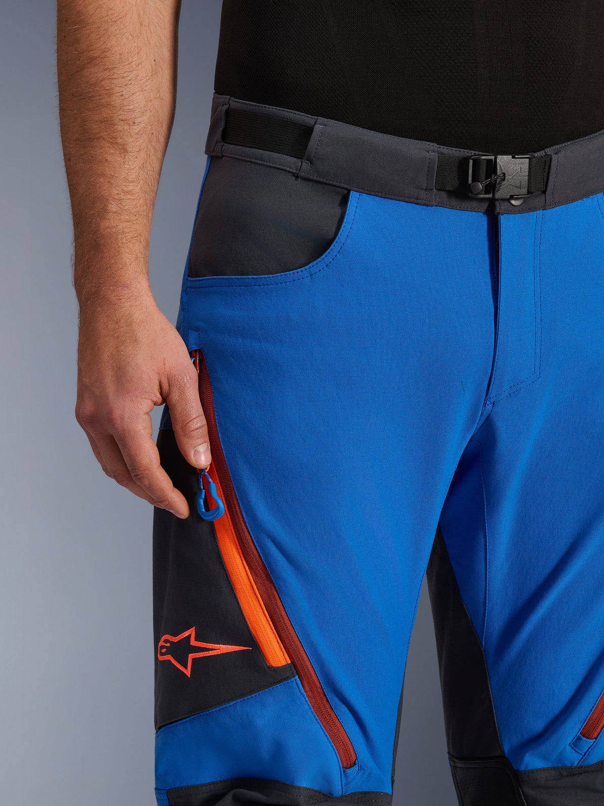 Calças Alpinestars Maxdura Dual em Azul Royal e Cinzento Escuro, vista de pormenor do cós com fivela magnética, bolso cargo com fecho e detalhes em laranja, logótipo Alpinestars vermelho, equipamento técnico de motociclismo de aventura