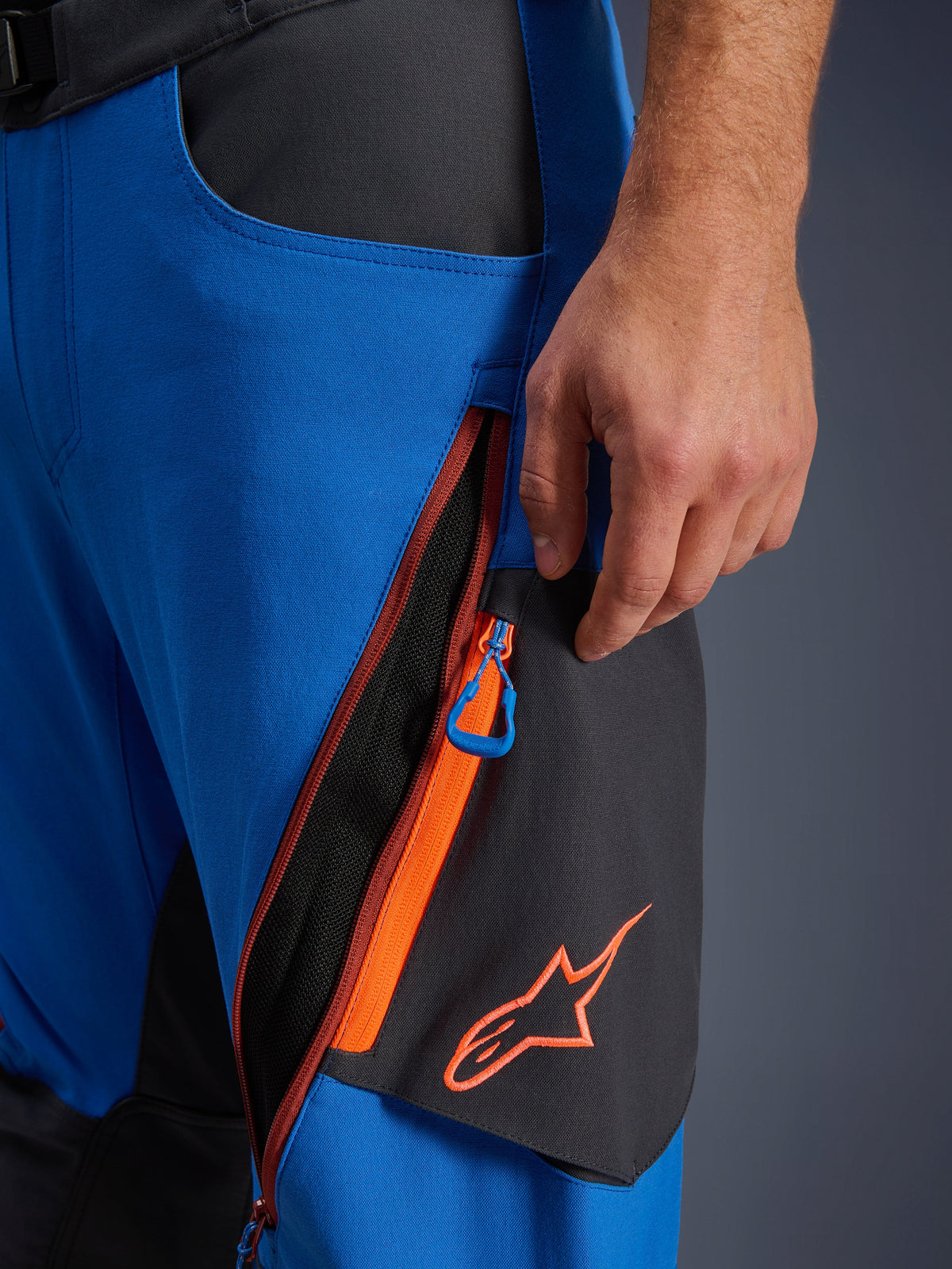 Detalhe das calças Alpinestars Maxdura Dual, calças de motociclismo de aventura em azul royal e cinzento escuro, grande plano da ventilação com fecho na coxa com detalhes cor de laranja e puxador azul, logótipo Alpinestars cor de laranja em painel preto, construção durável em poliamida elástica