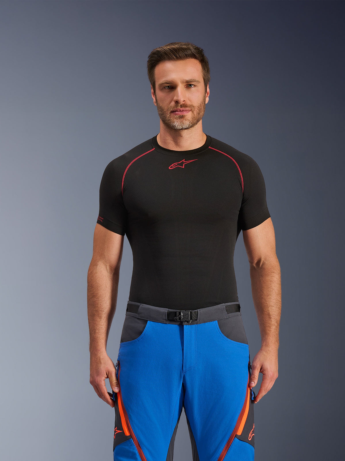 Calças Alpinestars Maxdura Dual, azul real e cinzento escuro, calças de motociclismo de aventura com detalhes em laranja, usadas por um modelo com uma camisola de compressão preta, com cinto de fivela magnética e aberturas de ventilação com fecho nas coxas, vestuário profissional de desportos motorizados