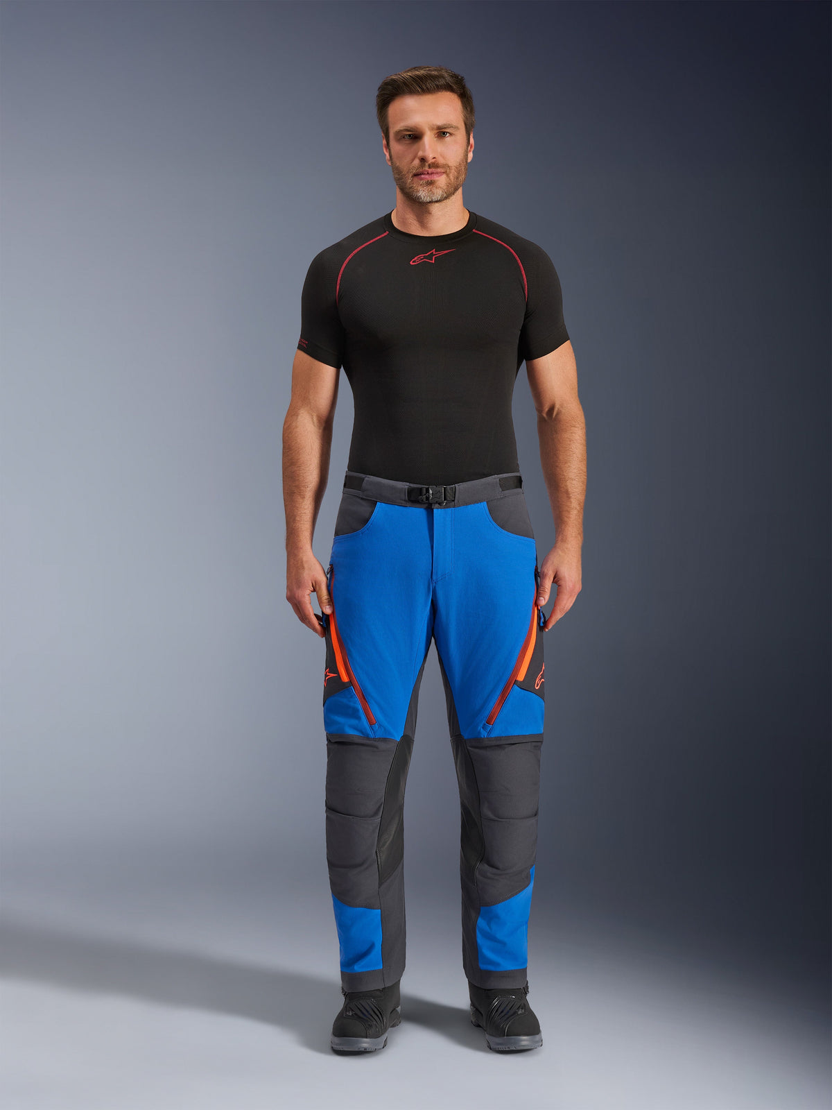 Alpinestars Maxdura Dual Pants, calças de motociclismo de aventura, azul royal e cinzento escuro, detalhes de fechos em laranja, usadas com uma camada de base Alpinestars preta e botas pretas, com painéis reforçados nos joelhos e design técnico off-road