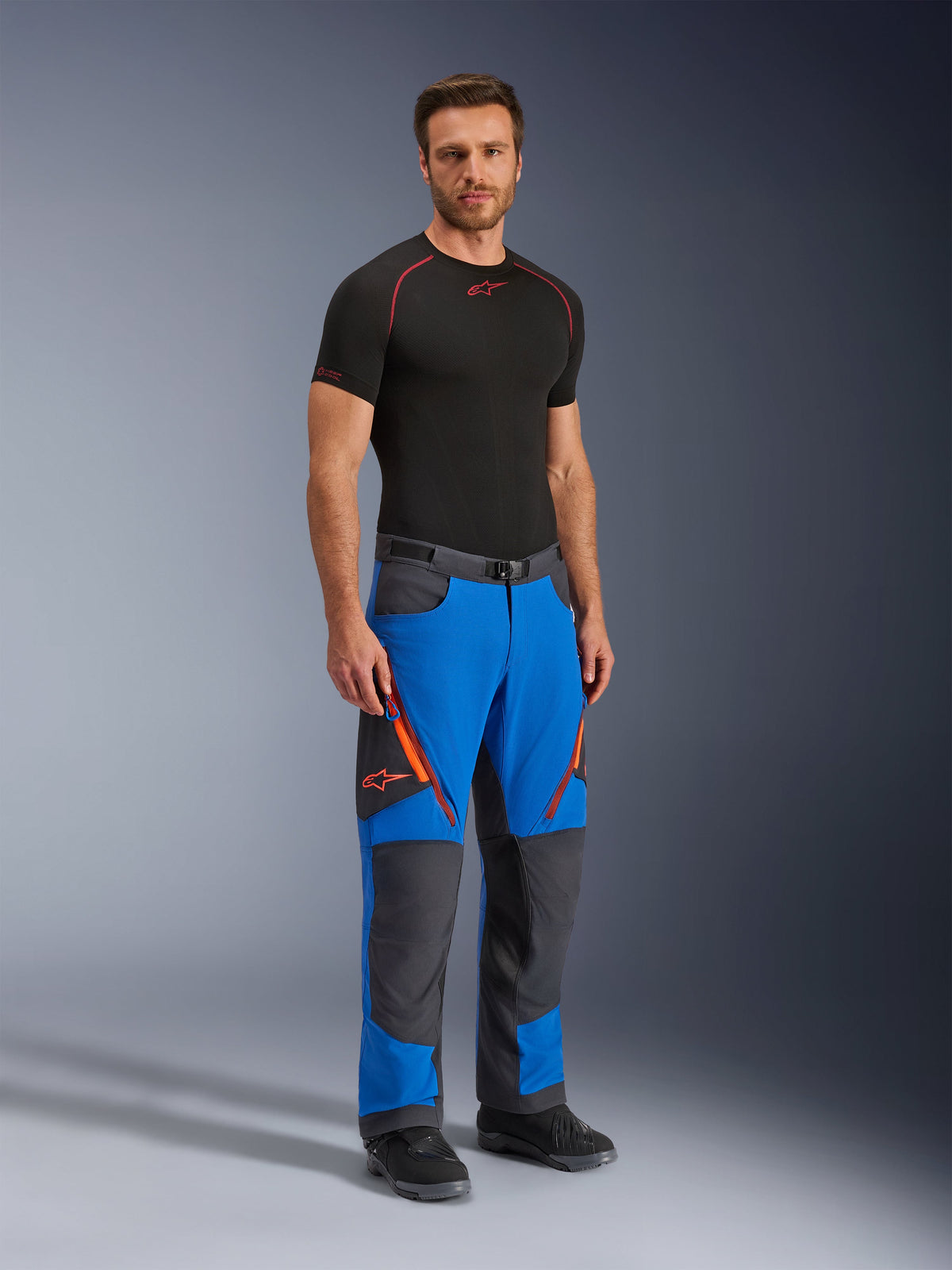 Alpinestars Maxdura Dual Pants, calças de motociclismo de aventura, azul royal e cinzento escuro, com detalhes de fecho e logótipos cor de laranja, vestidas por um modelo com camisola interior e botas pretas, design técnico com painéis reforçados para condução dual-sport