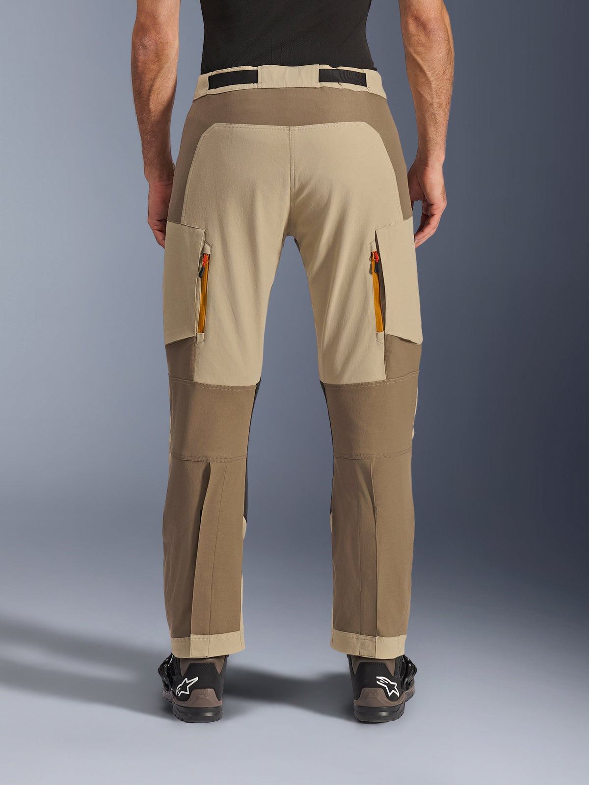 Alpinestars Maxdura Dual Pants, Walnut Aluminum, vista traseira de calças de motociclismo de aventura com painéis bege e em tons de terra, saídas de ventilação com fechos laranja e design por cima da bota, usadas com botas Alpinestars pretas.