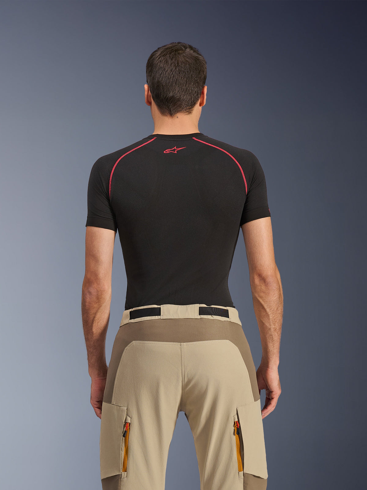 Calças Alpinestars Maxdura Dual, calças de motociclismo de aventura, Walnut Aluminum, vista traseira com painel de assento reforçado e tecido elástico bege, bolsos cargo com fecho de correr e puxadores cor-de-laranja, usadas com uma camisola base layer preta da Alpinestars.