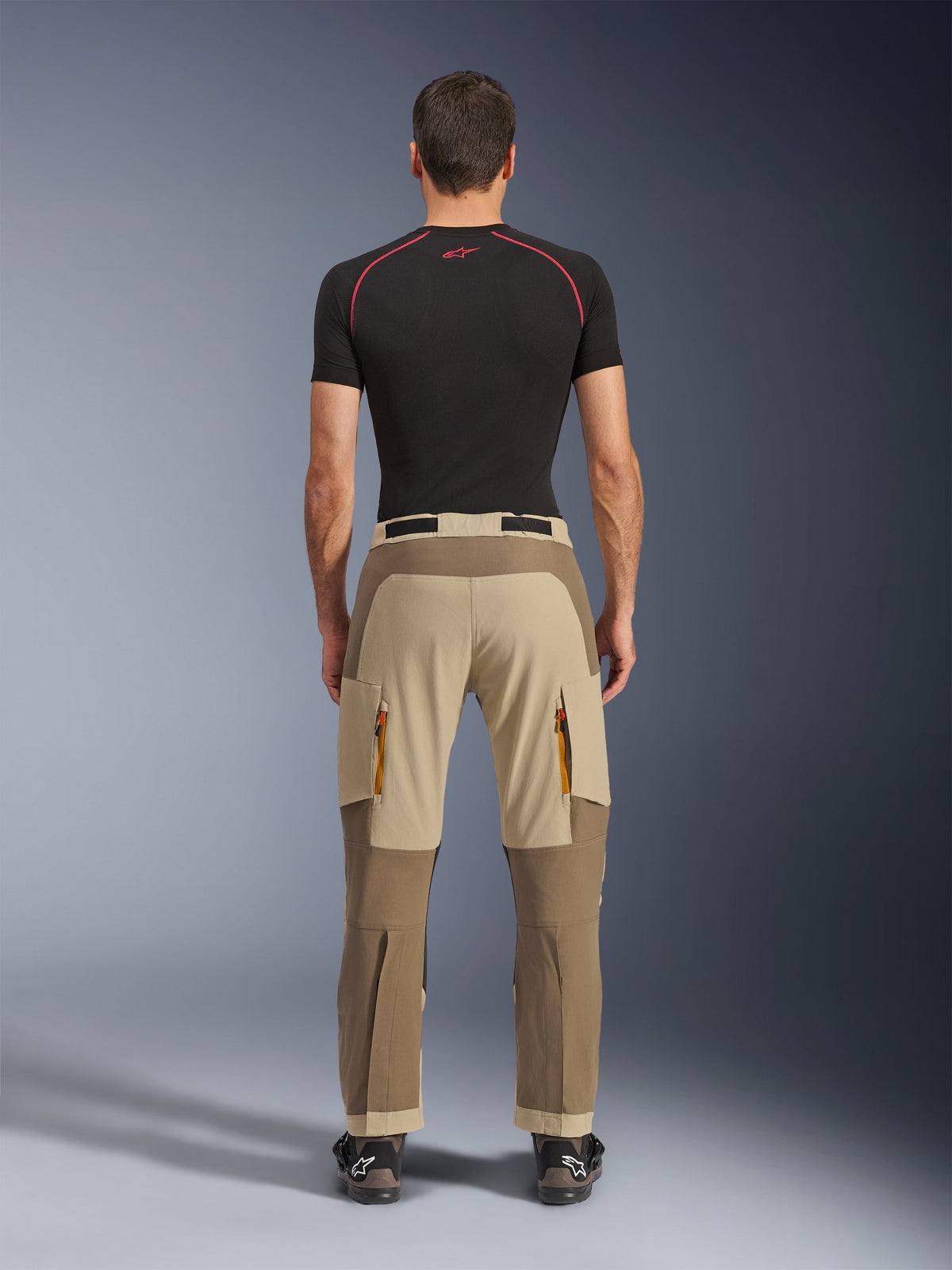 Alpinestars Maxdura Dual Pants em Walnut Aluminum, vista traseira com design em dois tons de castanho e bege, calças de aventura com bolsos cargo com fecho e assento reforçado, usadas com camisola de compressão preta e botas de motociclismo.