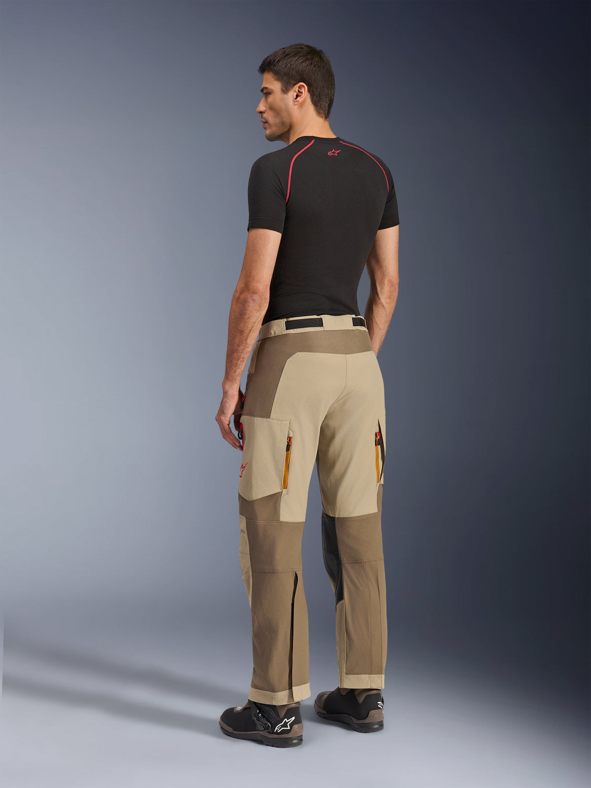 Alpinestars Maxdura Dual Pants, calças de condução de aventura e dual-sport, cor Walnut Aluminum, vista traseira num modelo com camisola interior preta e botas de condução, com construção multi-painel, aberturas traseiras com fecho e reforços sobrepostos.