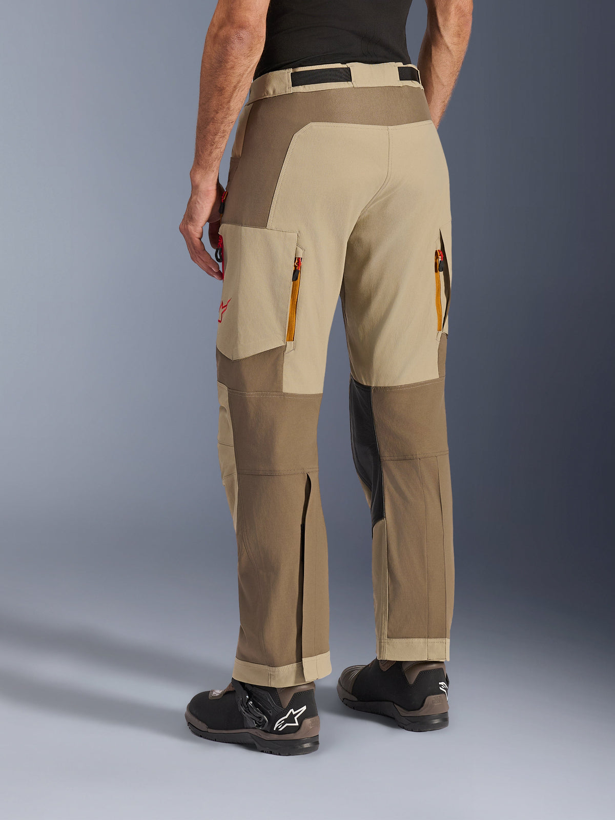 Alpinestars Maxdura Dual Pants em Walnut Aluminum, vista lateral traseira, calças de motociclismo de aventura com painéis bege e castanhos, saídas de ventilação traseiras com fecho éclair, bolsos cargo grandes e design por cima da bota, usadas com botas pretas e castanhas